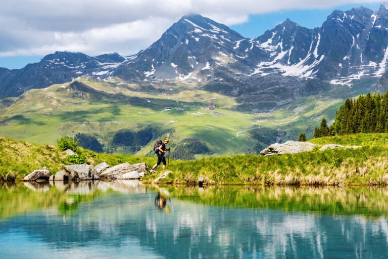 Female traveler with backpack hiking mountain trail next to a mountain lake and admiring views of Swiss Alps in Val de Bagnes area, Switzerland.